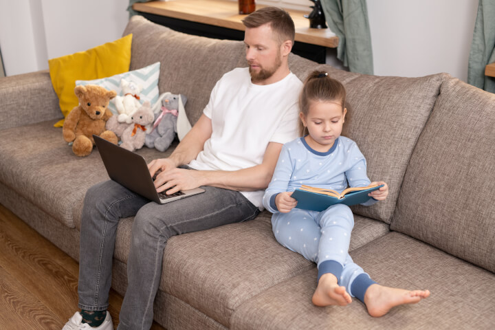 pria di laptop dengan putrinya membaca di sofa